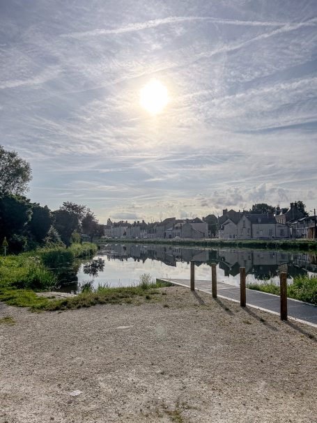 Sonnenstrahlen über einem ruhigen Gewässer, umgeben von Bäumen und Bauten im Hintergrund.