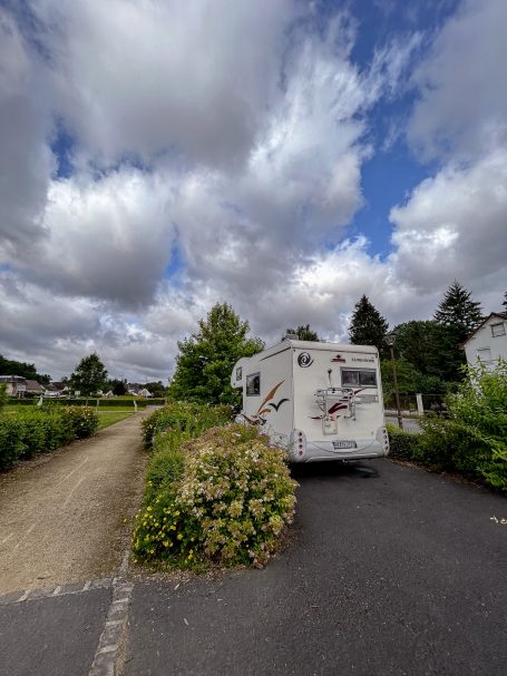 Wohnmobil auf einem Platz, umgeben von Bäumen und bewölktem Himmel.