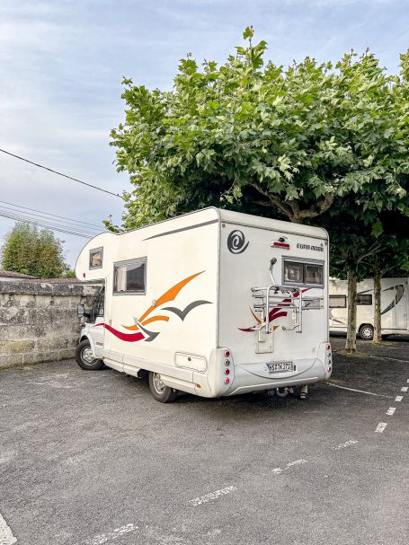 Weißer Wohnmobil parkt unter Bäumen auf einem Parkplatz.